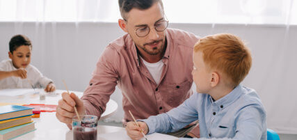 teacher helping little boy to draw picture at table in classroom