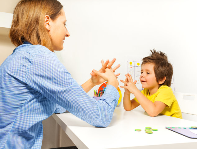 Boy exercises by putting hands and fingers together with therapist showing it improving motor skills sitting opposite at the table indoors