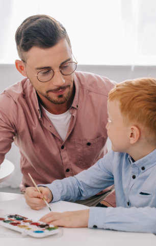 teacher helping little boy to draw picture at table in classroom