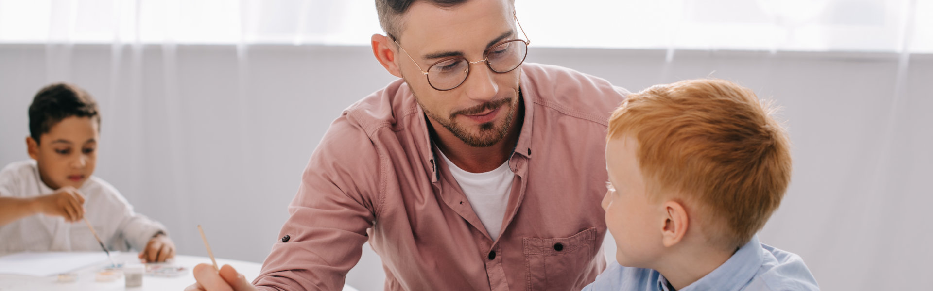 teacher helping little boy to draw picture at table in classroom